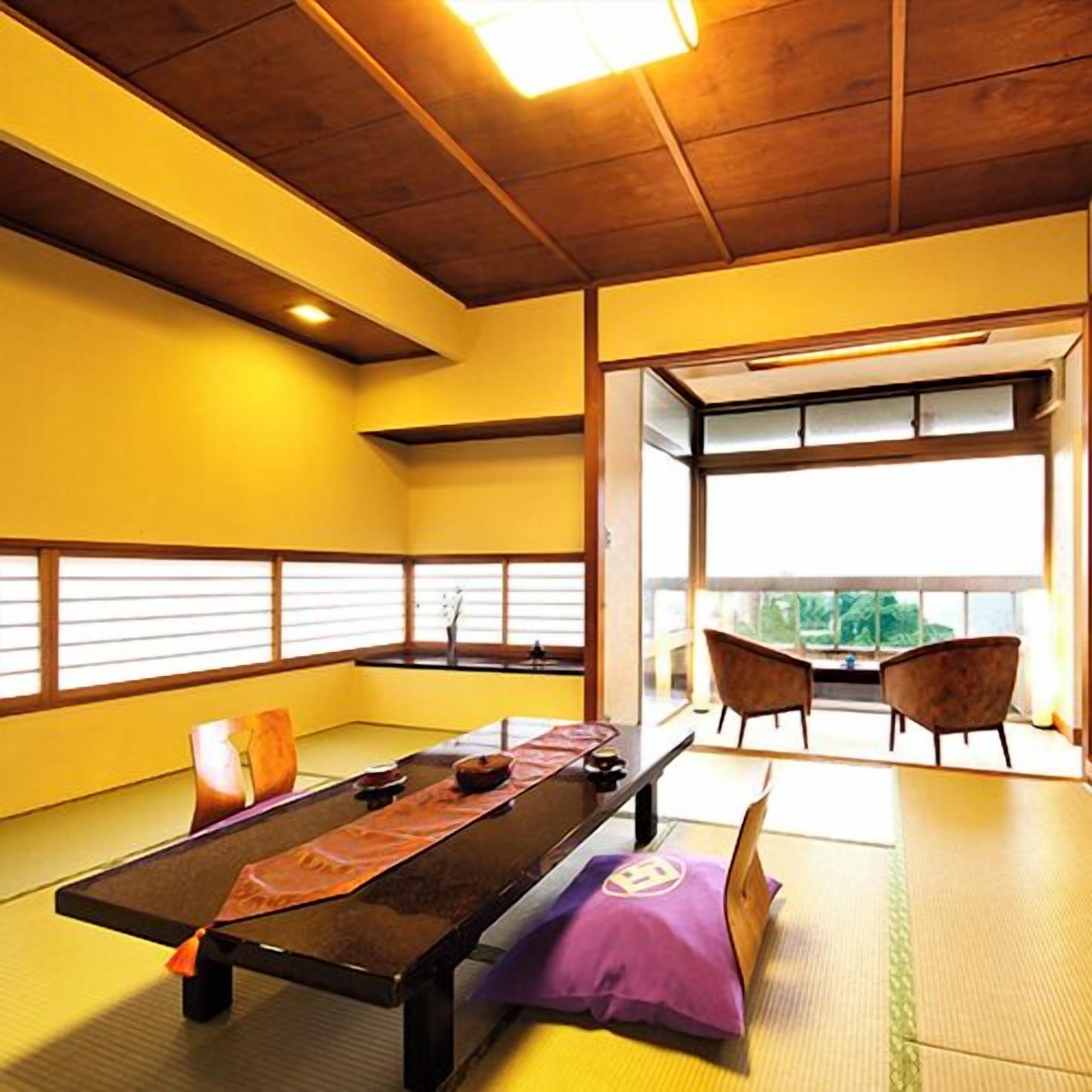 Ein Raum mit Tatami-Matten, einem niedrigen Tisch, Kissen und Blick durch ein großes Fenster.