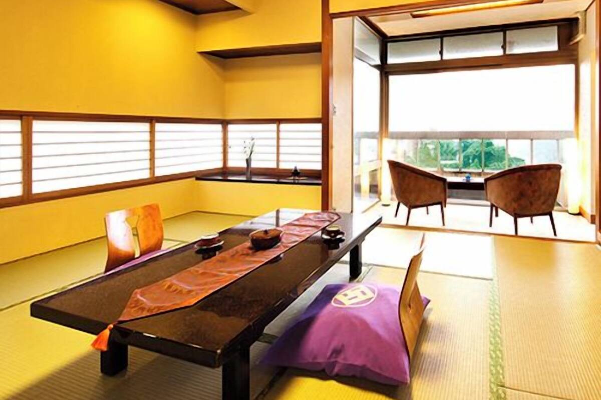 A tatami mat room with a low table, cushions, and a view through a large window.