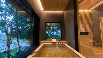 A modern spa with a large rectangular hot tub, wooden ceiling, and large windows offering a view of trees.