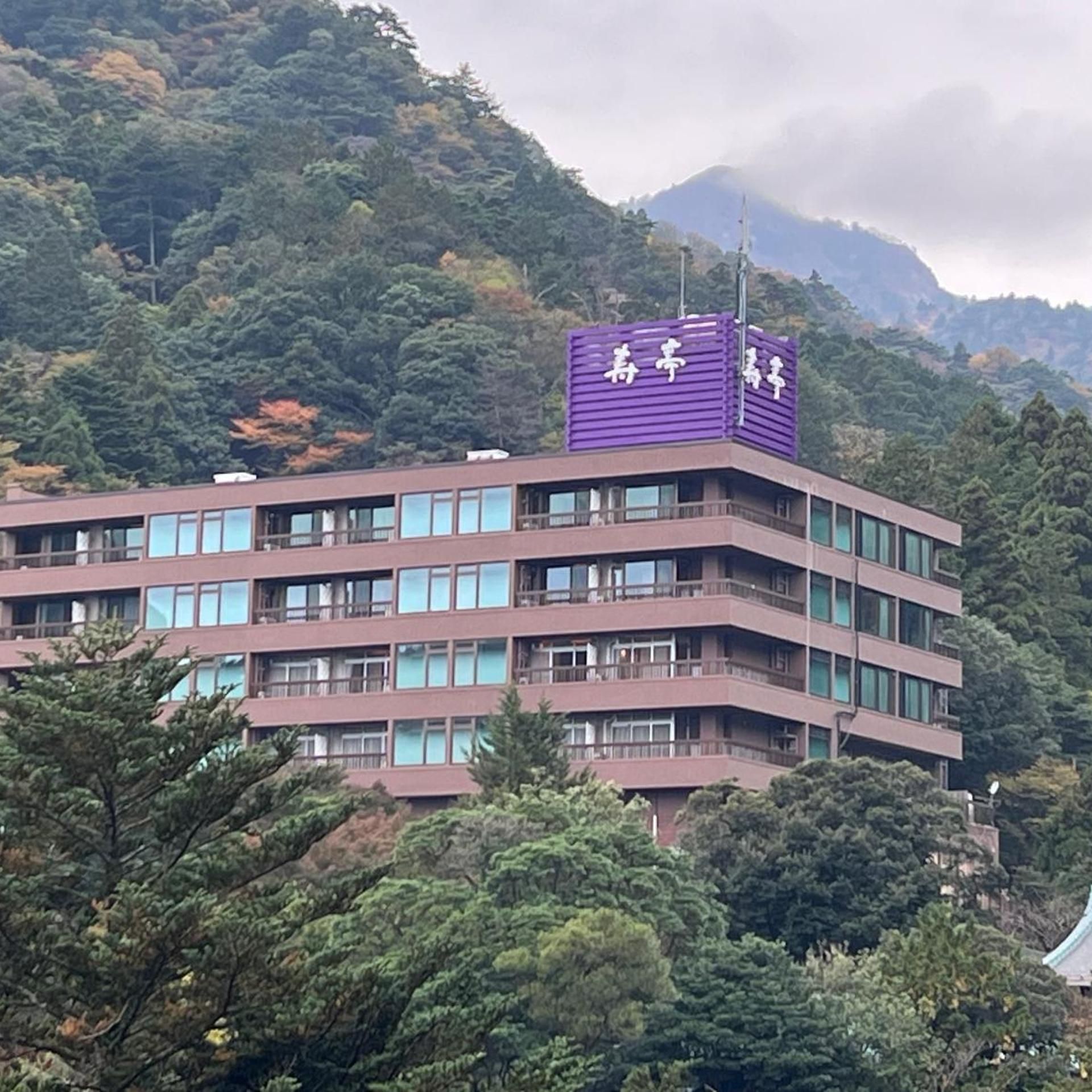 A multi-story building with a purple sign on top, surrounded by dense greenery and a mountain in the background.