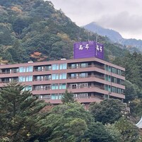 Un edificio de varios pisos con un letrero púrpura en la parte superior, rodeado de abundante follaje y una montaña al fondo.