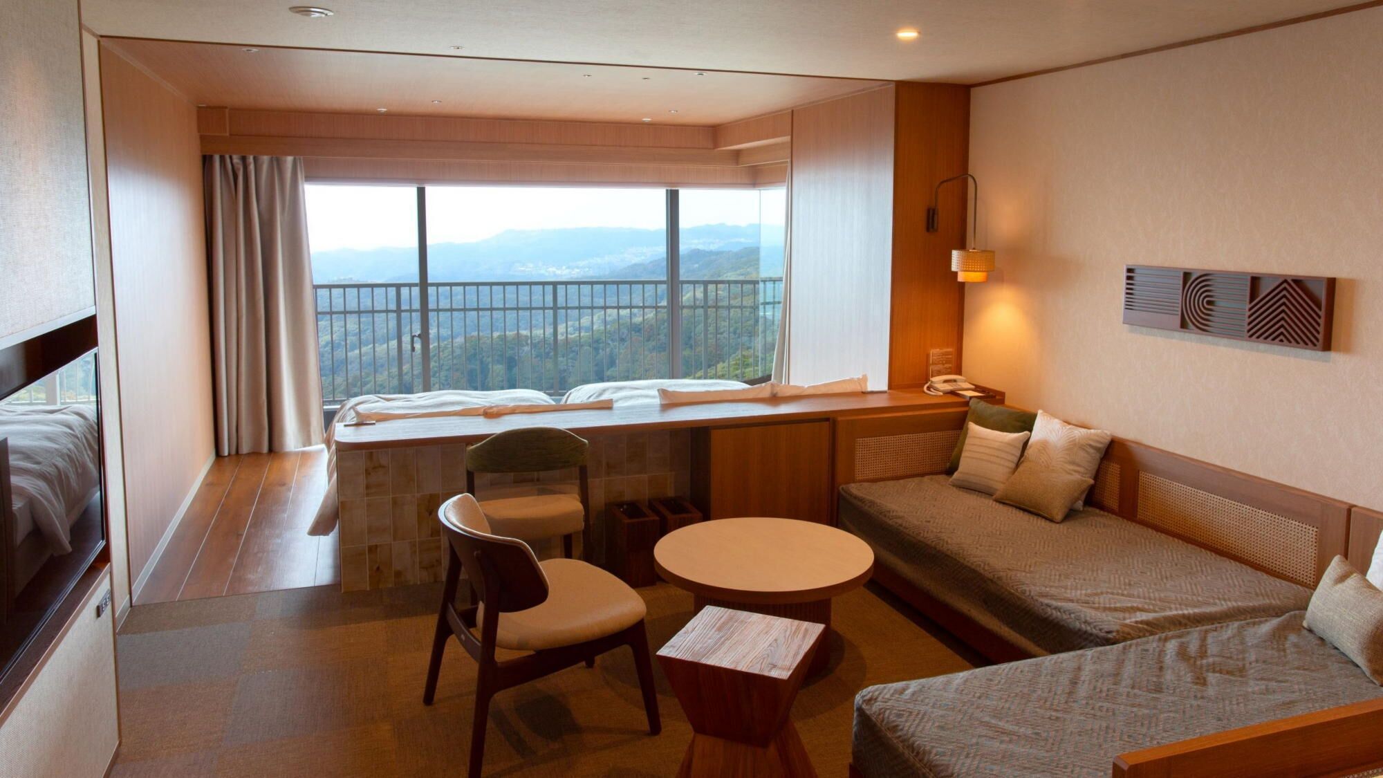 A hotel room with a bed, a sofa, a desk, a chair, a small table, a window with a view, and a wall-mounted phone.
