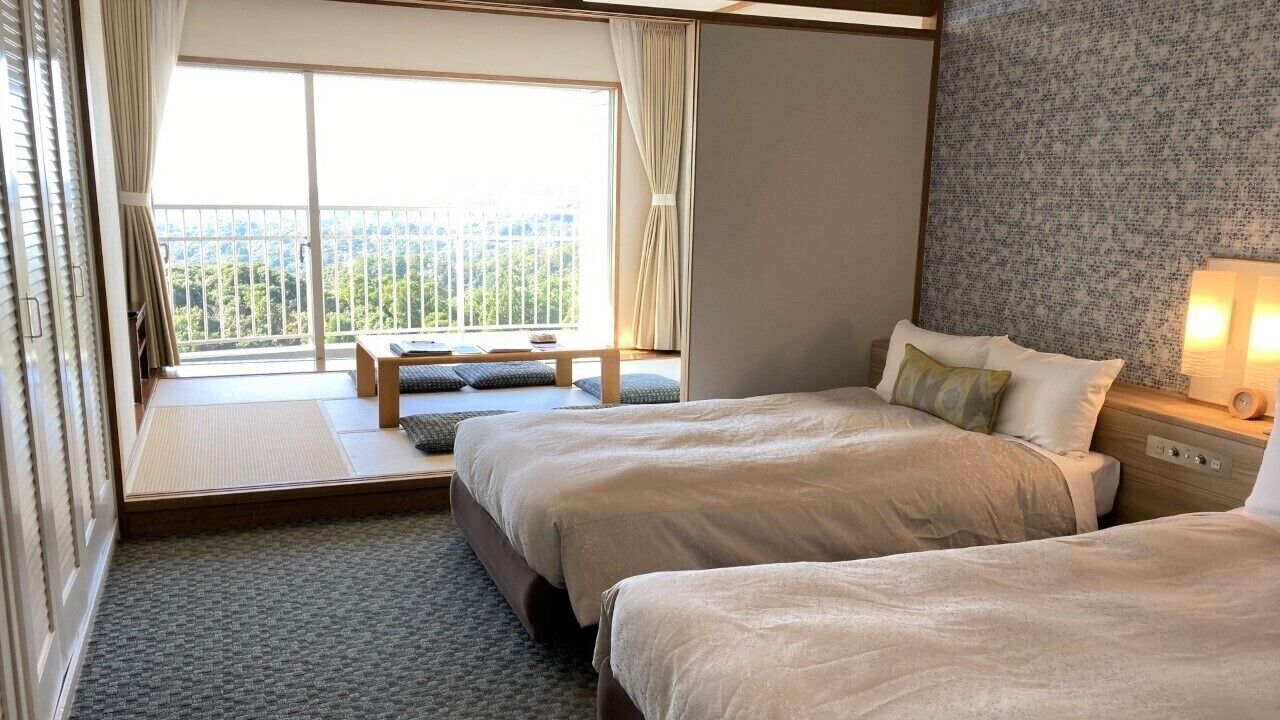 A hotel room with two beds, a window with a view, a bedside table with a lamp, and a small bench by the window.