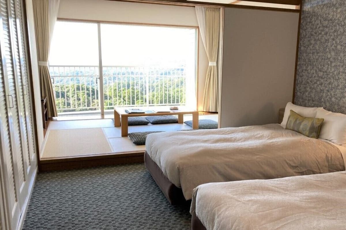 Ein Hotelzimmer mit zwei Betten, einem Fenster mit Blick, einem Nachttisch mit Lampe und einer kleinen Bank am Fenster.