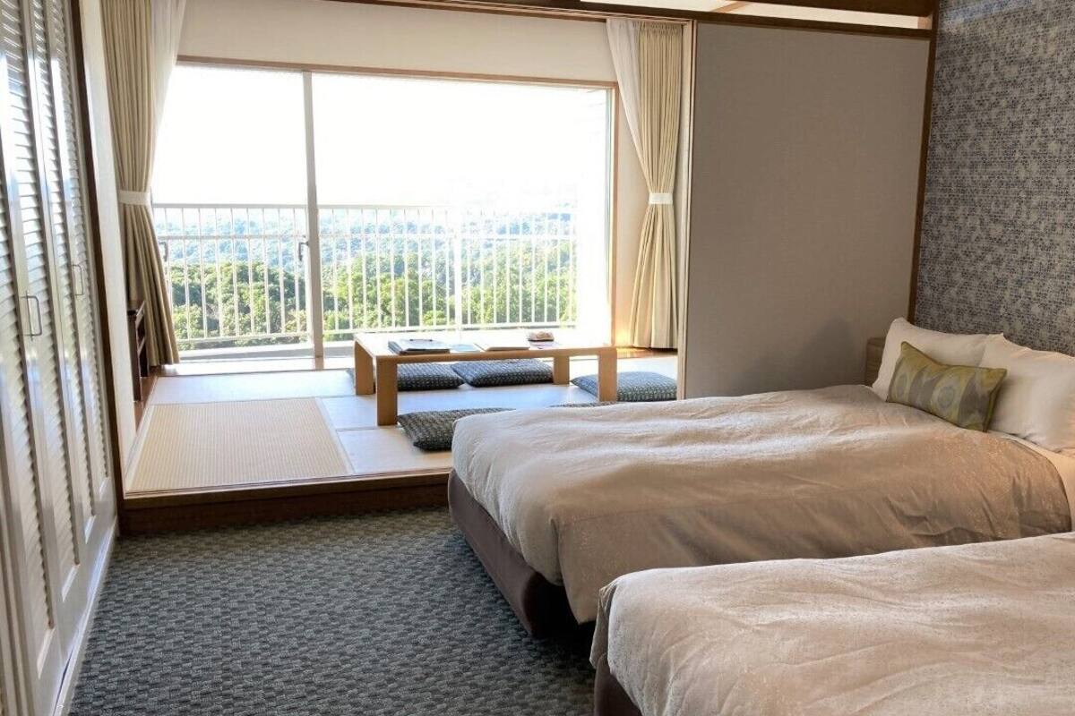 Habitación de hotel con dos camas, una ventana con vistas, una mesita de noche con lámpara y un pequeño banco junto a la ventana.