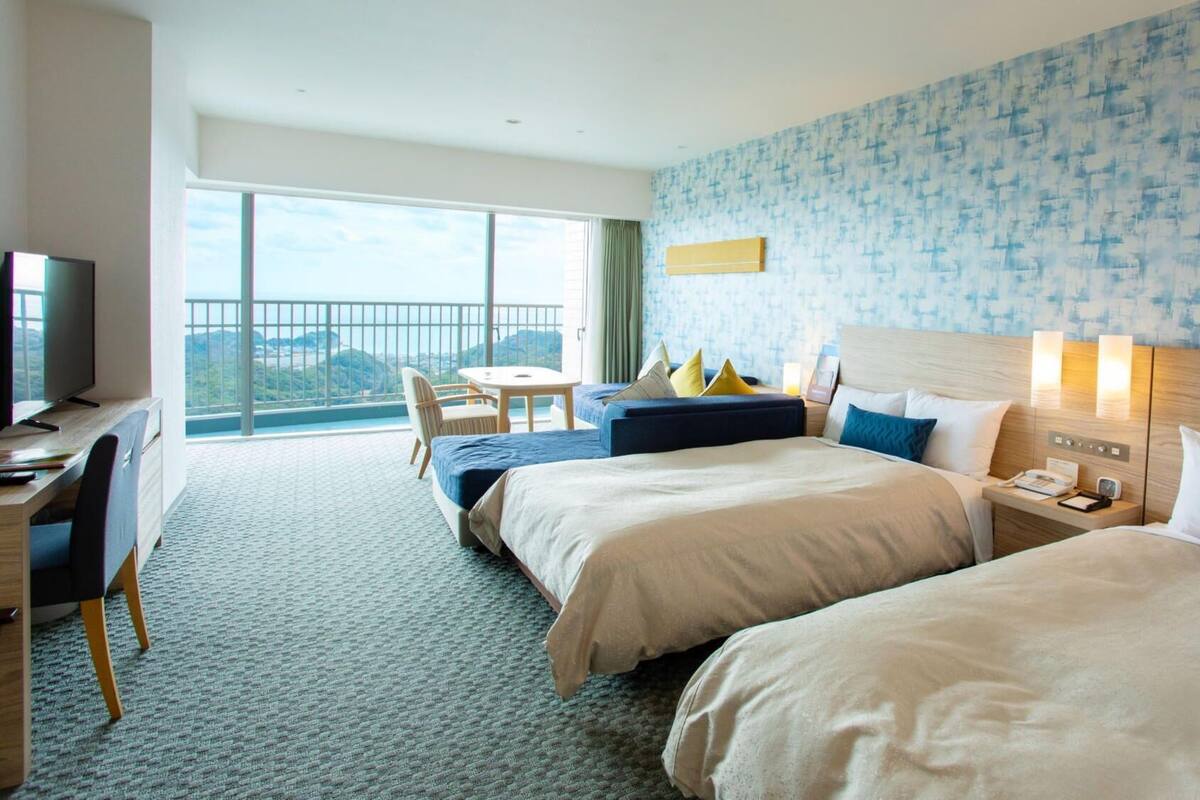 Ein Hotelzimmer mit zwei Betten, einem Schreibtisch, einem Stuhl, einem Fernseher und einem Balkon mit Blick.