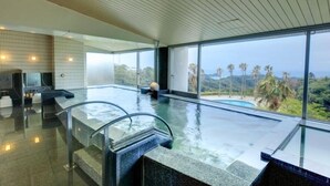 A modern indoor pool with clear glass walls, a stone floor, and a view of palm trees and hills. - Hotel Harvest Katsuura (Katsuura)