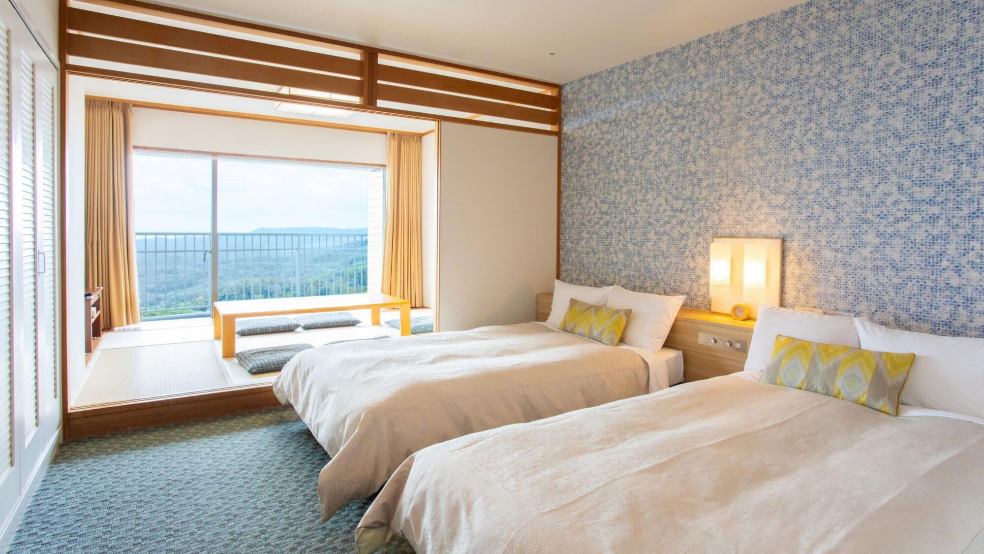 A hotel room with two beds, a large window with a view, a wooden bench, and a patterned wall.