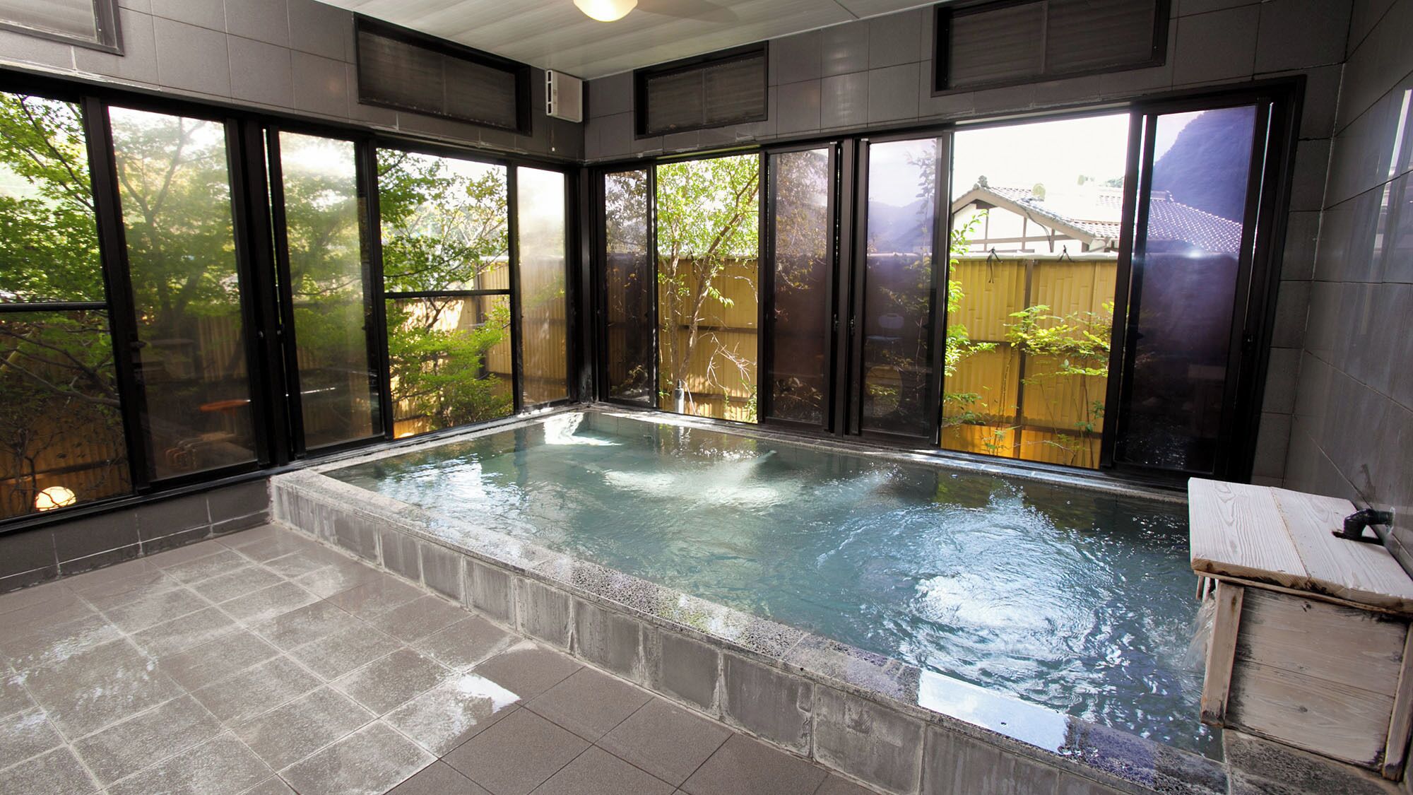 A spa-like room with a hot tub, large windows, and a view of a wooden fence and trees.
