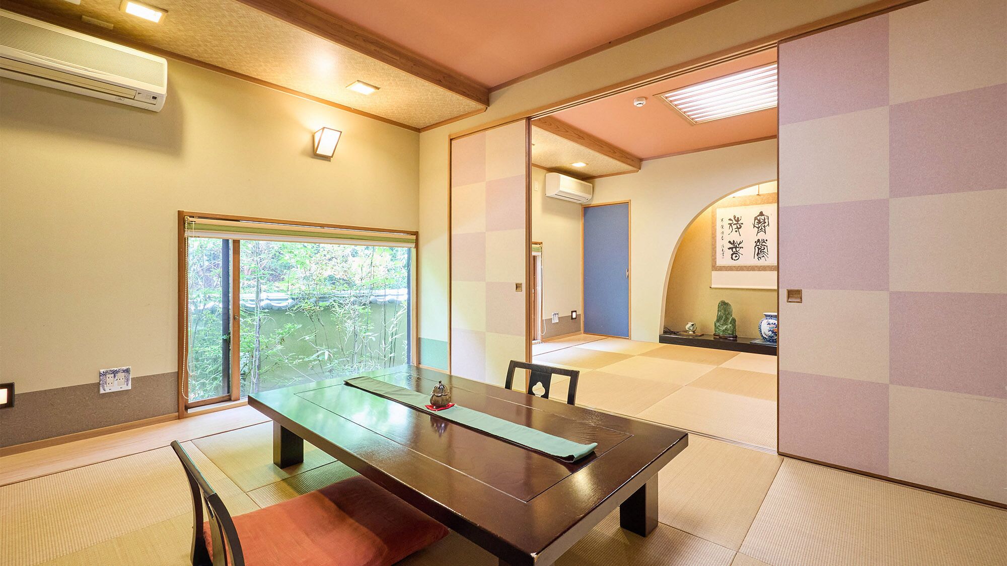 Ein traditioneller japanischer Raum mit Tatami-Böden, einer Schiebetür, einem Tisch mit Unterlage und Blick ins Grüne.