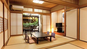 Een traditionele Japanse kamer met tatami vloeren, een lage tafel, stoelen, een televisie en een schuifdeur die uitkomt op een tuin.