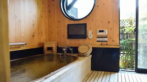 Een houten sauna met een groot bad, een klein krukje, een ronde spiegel en een bedieningspaneel.