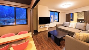 Un salon moderne avec un canapé, une table basse, un téléviseur et une vue sur une forêt, visible à travers de grandes fenêtres.