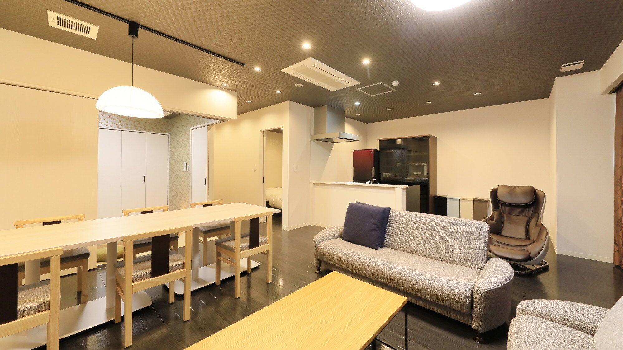 A modern living room with a sofa, a massage chair, a dining table with chairs, and a kitchen area with cabinets and a refrigerator.
