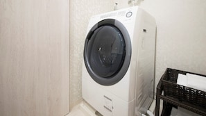 A compact, white front-loading washing machine in a small laundry area with a wicker basket containing folded towels. - Point Vacation Kaga Yamanaka (Kaga)