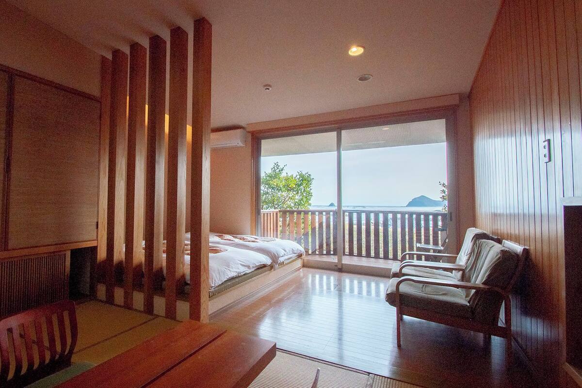 A room with a bed, a chair, a table, and a balcony with a view.