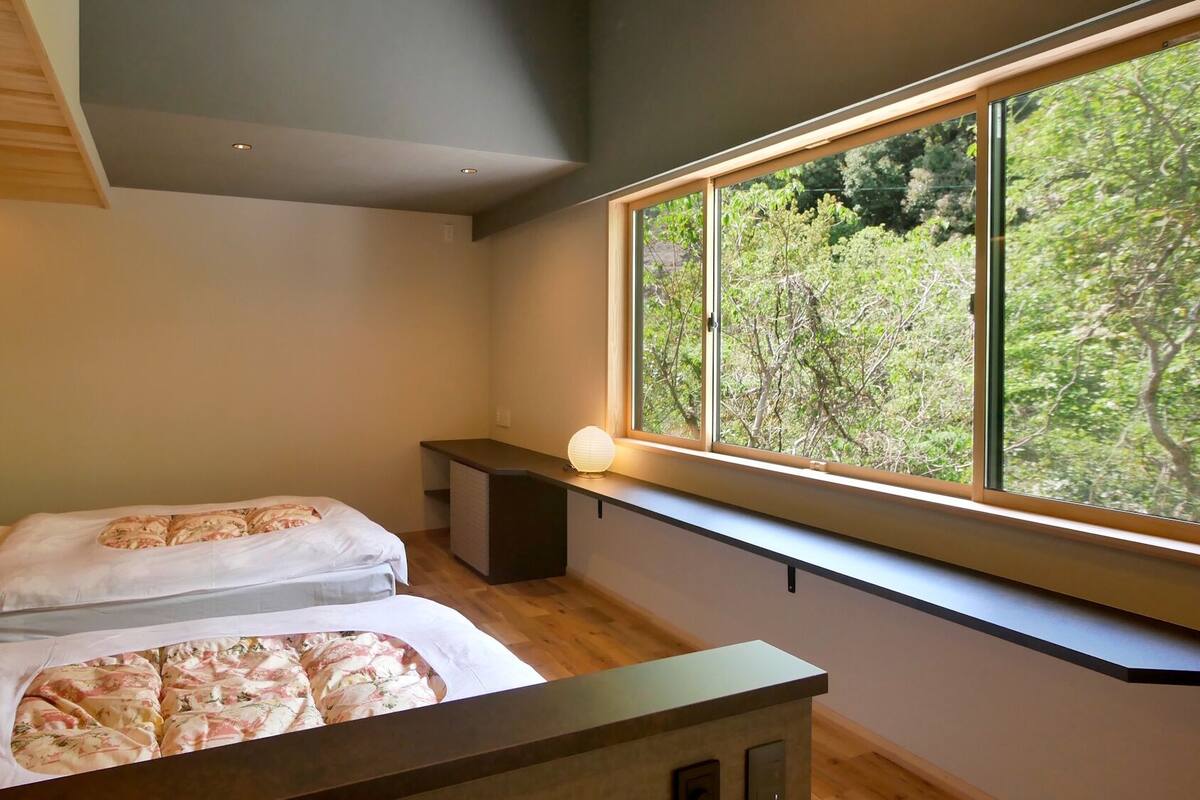 A bedroom with two beds, a bedside table, a window with a view of trees, and a ceiling light.