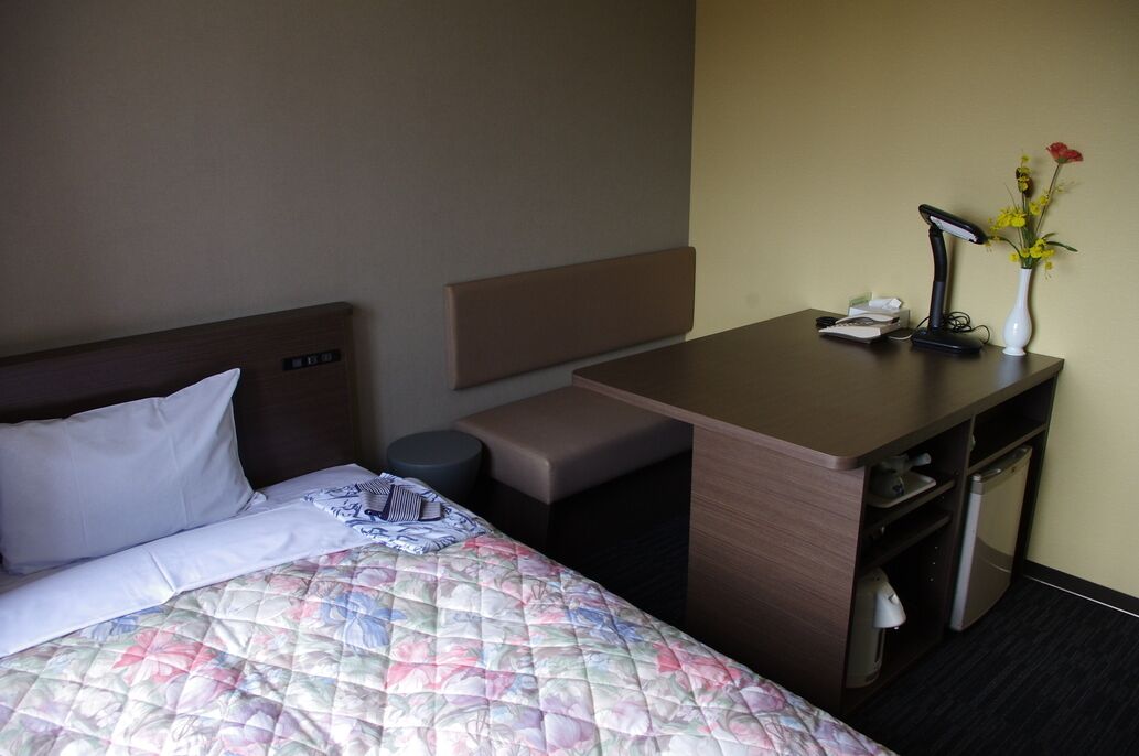 A hotel room with a bed, a desk, a chair, a small trash bin, a lamp, and a vase with flowers.