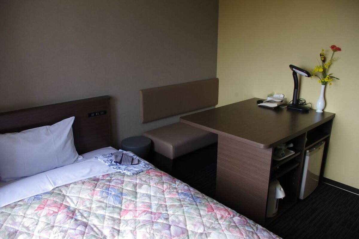 A hotel room with a bed, a desk, a chair, a small trash bin, a lamp, and a vase with flowers.