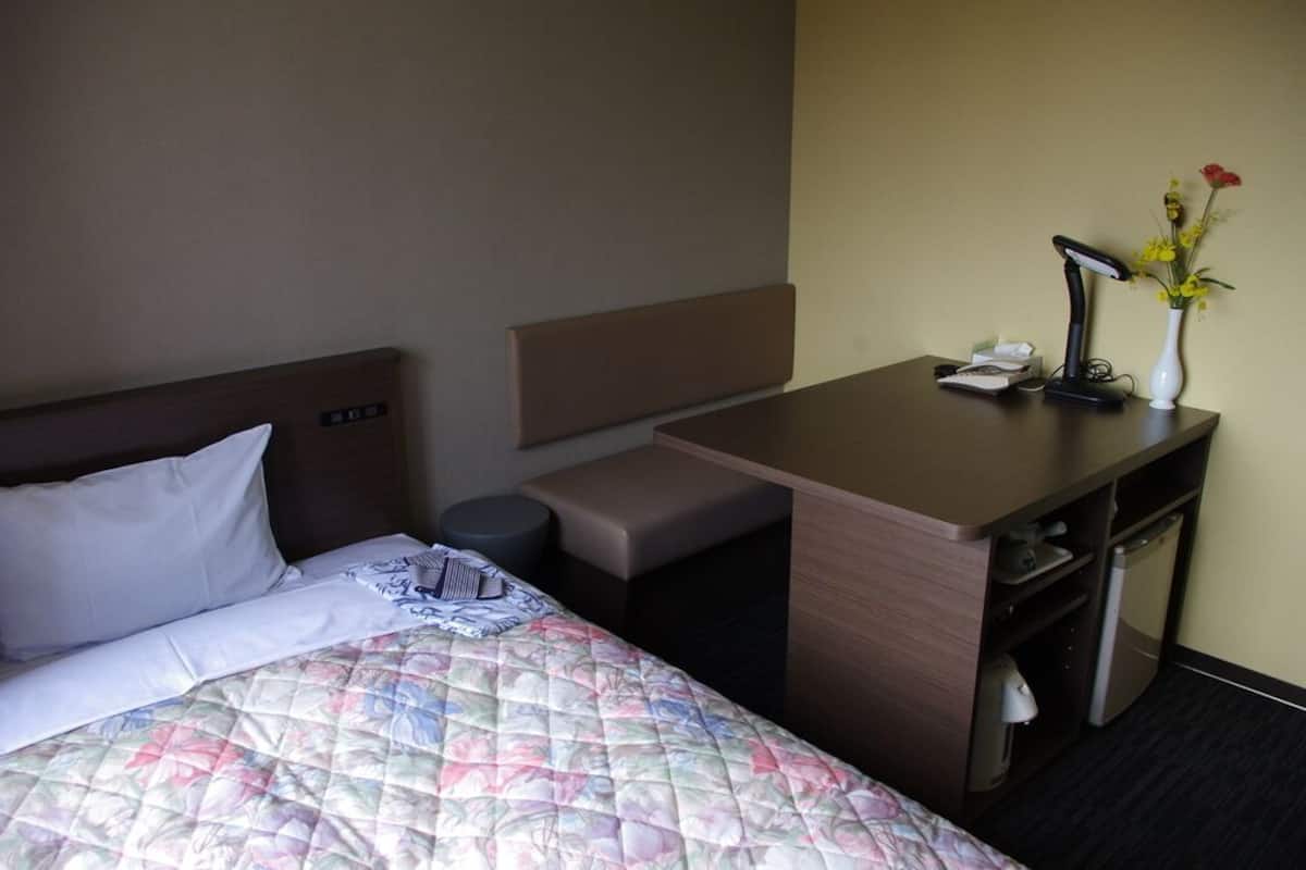 Quarto de hotel com cama, escrivaninha, cadeira, lixeira pequena, abajur e um vaso com flores.