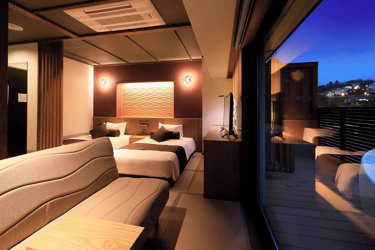 Une chambre d’hôtel moderne avec un balcon, un lit doté de plusieurs oreillers, une table de chevet et une grande fenêtre.