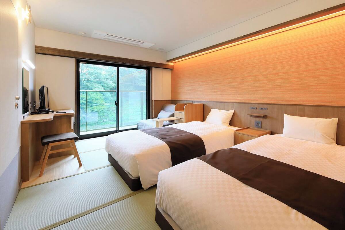 Une chambre d’hôtel avec deux lits, un bureau avec un ordinateur, une chaise, une télévision et une porte-fenêtre coulissante donnant sur l’extérieur.
