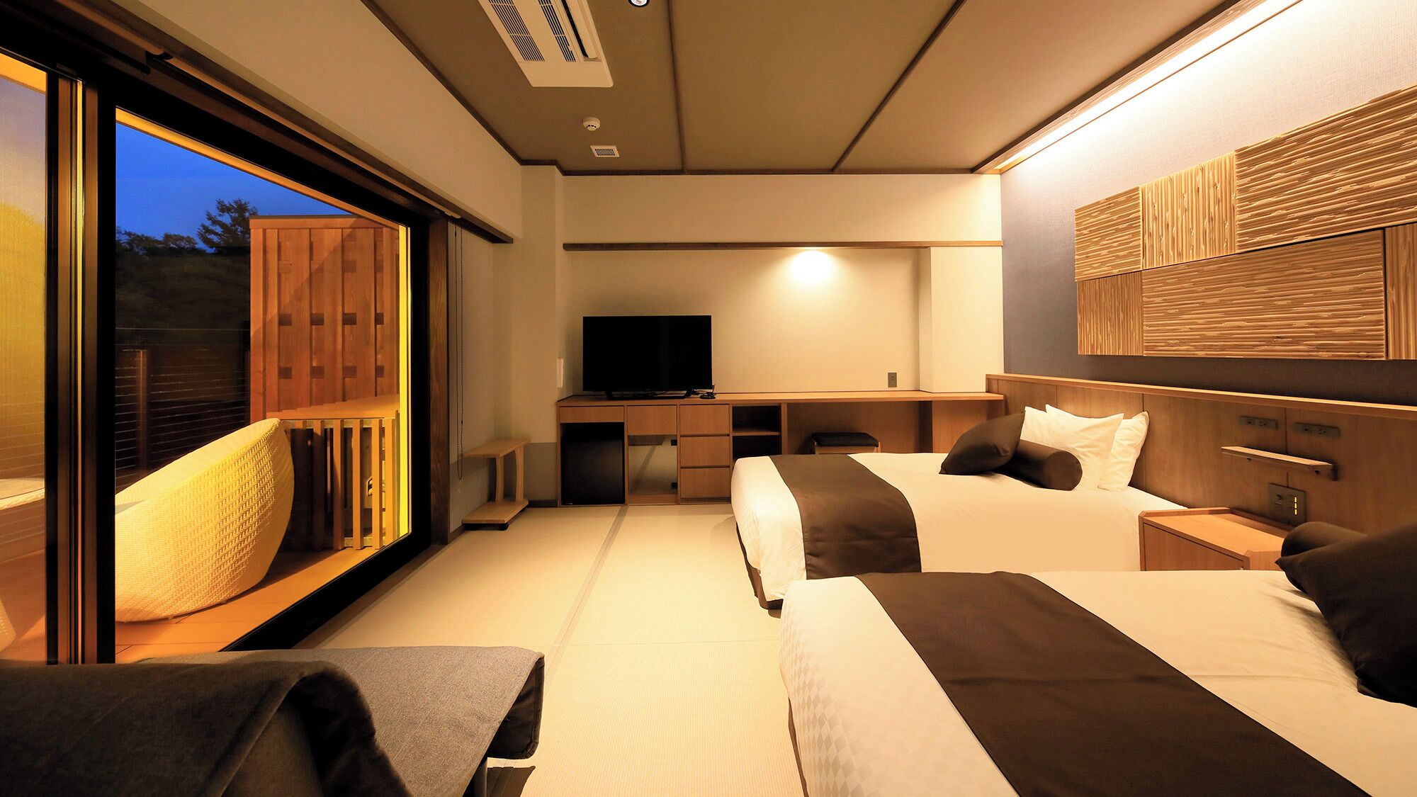A hotel room with a large window, a desk with a television, two beds, and a wooden headboard.