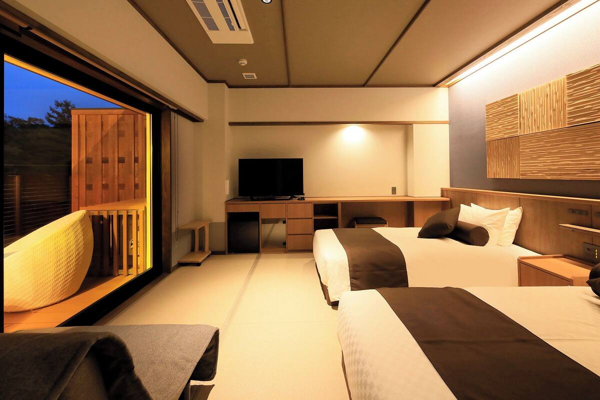 Une chambre d’hôtel avec une grande fenêtre, un bureau avec une télévision, deux lits et une tête de lit en bois.