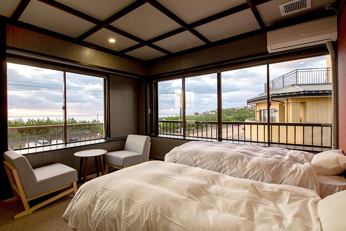 A hotel room with two beds, a small round table, a chair, and large windows offering a view of the city.