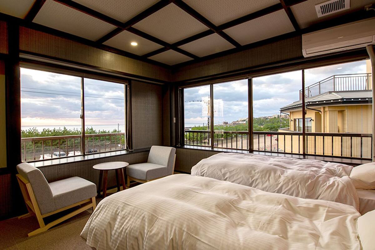 A hotel room with two beds, a chair, a small table, and large windows offering a view of the city.
