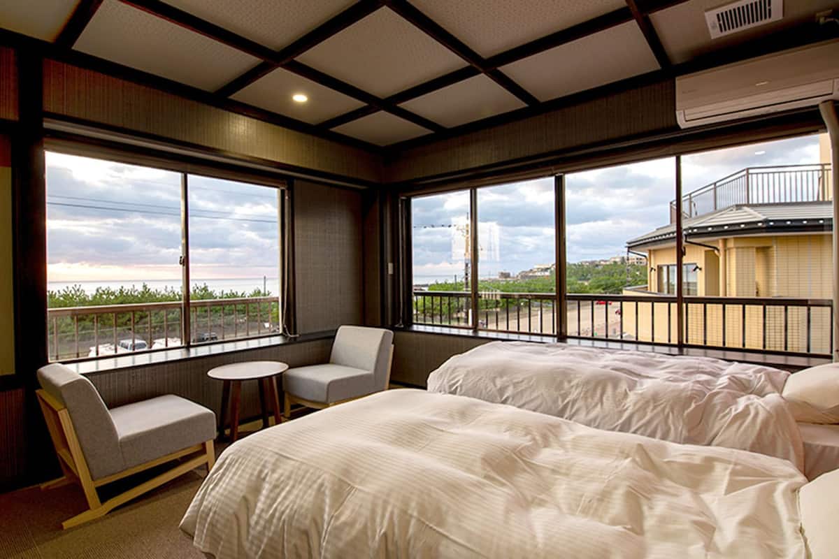 A hotel room with two beds, a chair, a small table, and large windows offering a view of the city.