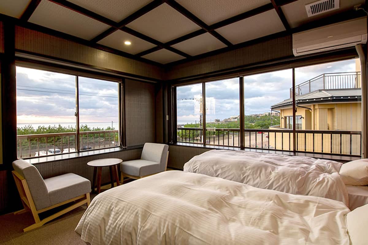 A hotel room with two beds, a small round table, a chair, and large windows offering a view of the city.