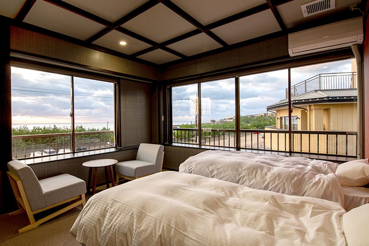 A hotel room with two beds, a chair, a small table, and large windows offering a view of the city.
