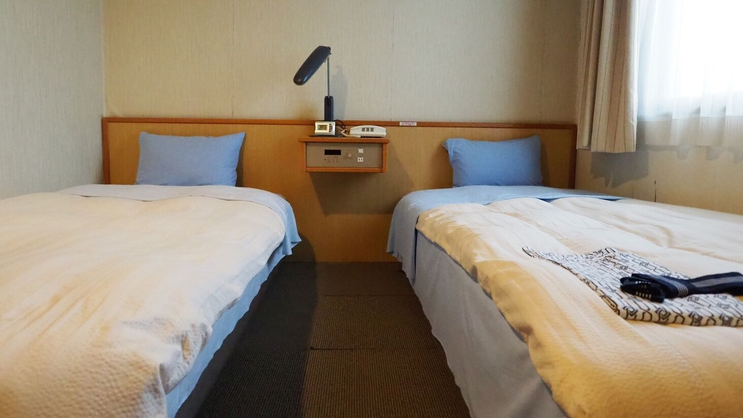 A hotel room with two beds, a nightstand with a radio and clock, and a window with curtains.