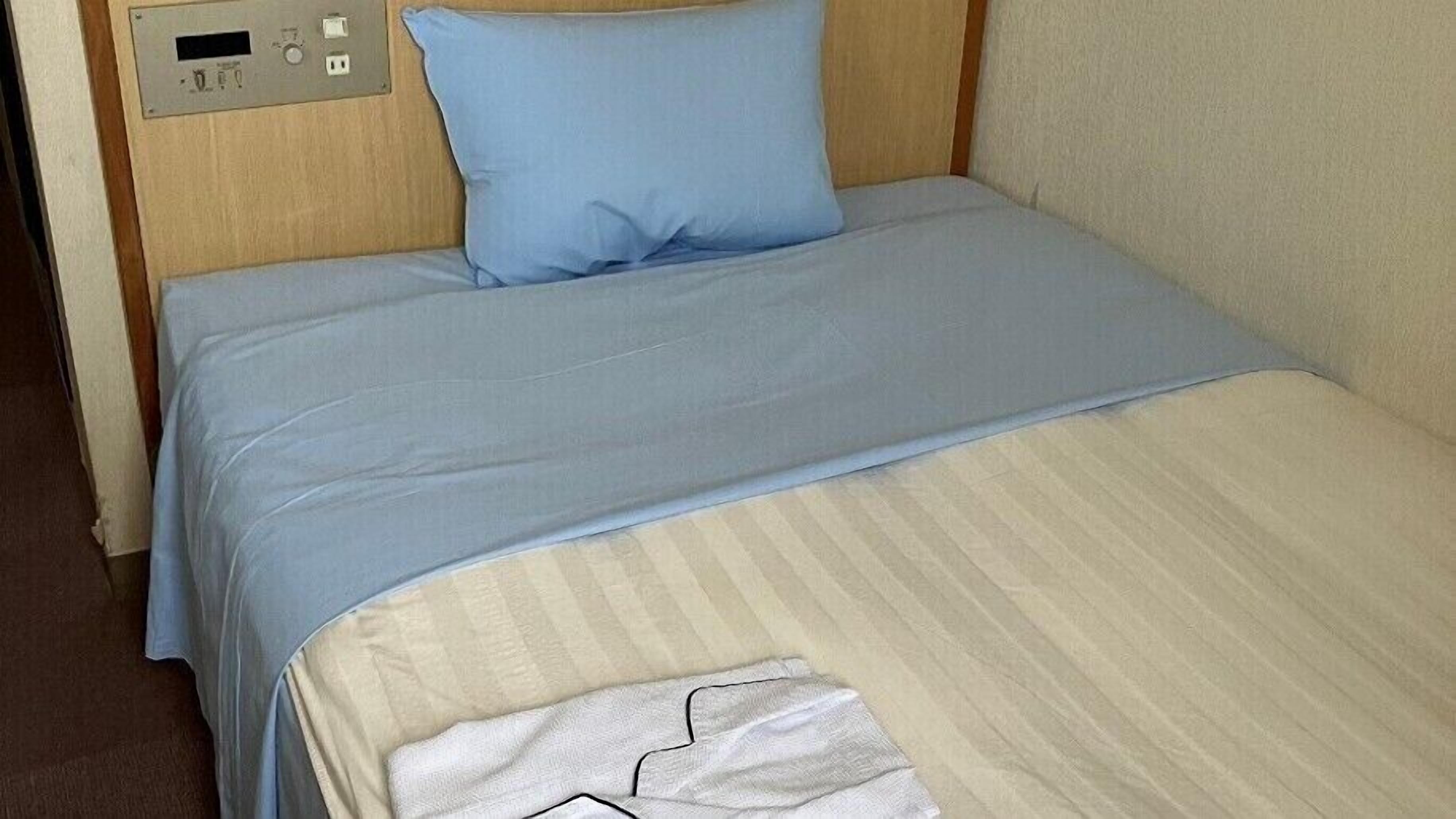 A bed with a blue pillow and a beige blanket, a control panel on the wall, and a white towel on the bed.