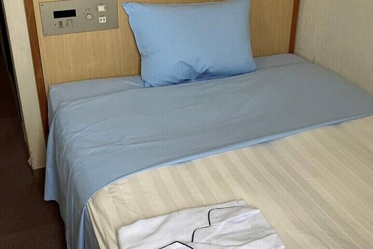 A bed with a blue pillow and a beige blanket, a control panel on the wall, and a white towel on the bed.