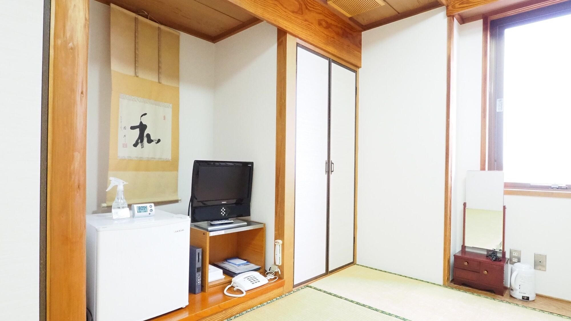 A room with a wooden wardrobe, a television on a stand, a small desk with a telephone, and a mirror.