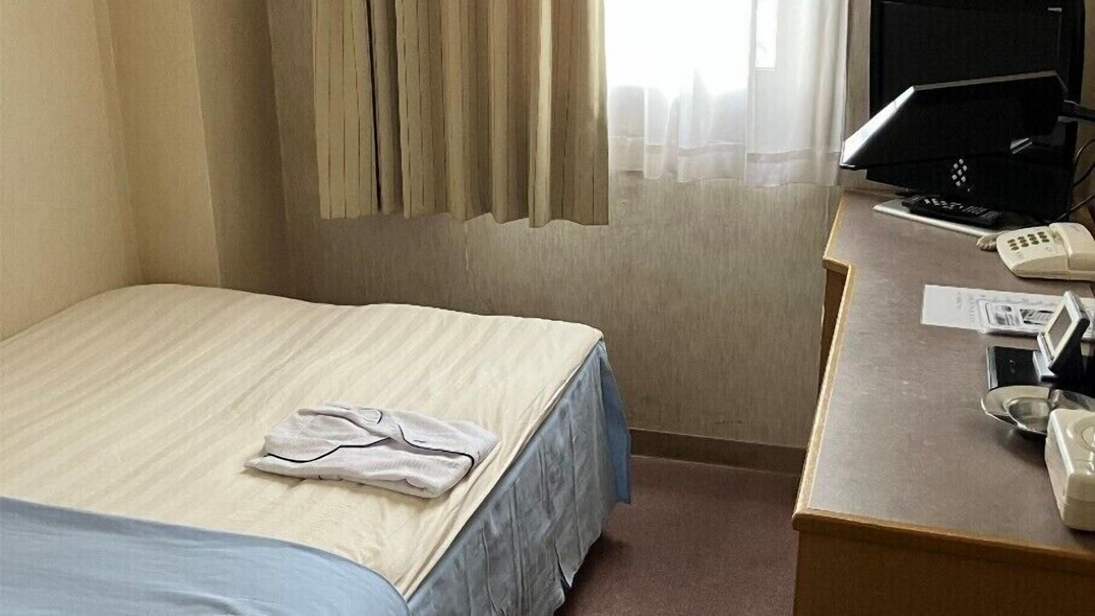 A hotel room with a bed, a desk with a computer and phone, and a window with curtains.