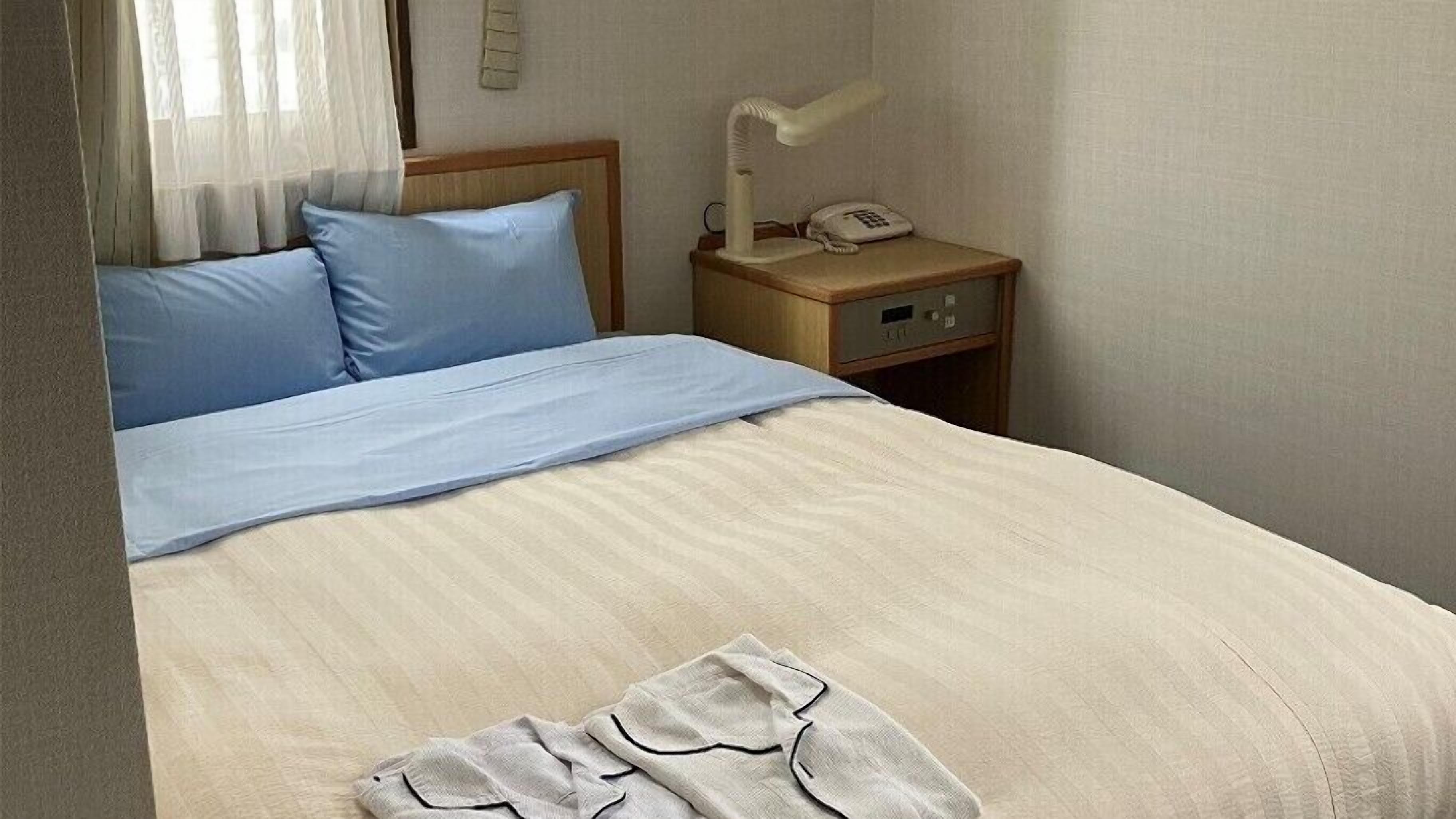 A bed with a blue pillow and a white blanket, a bedside table with a lamp and a phone, and a wooden headboard.