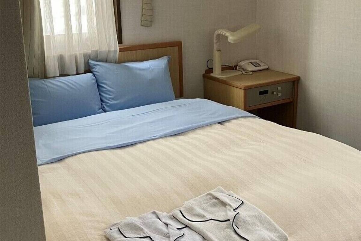 A bed with a blue pillow and a white blanket, a bedside table with a lamp and a phone, and a wooden headboard.