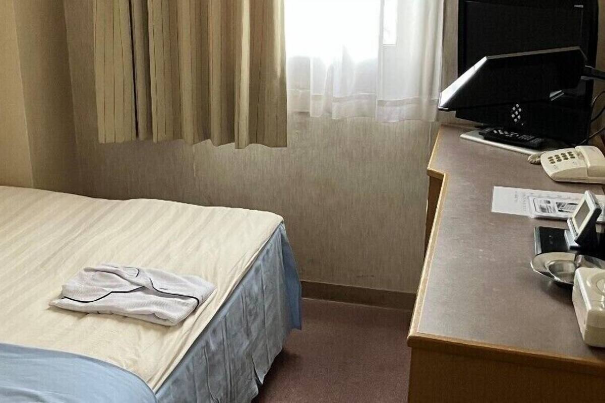 A hotel room with a bed, a desk with a computer and phone, and a window with curtains.