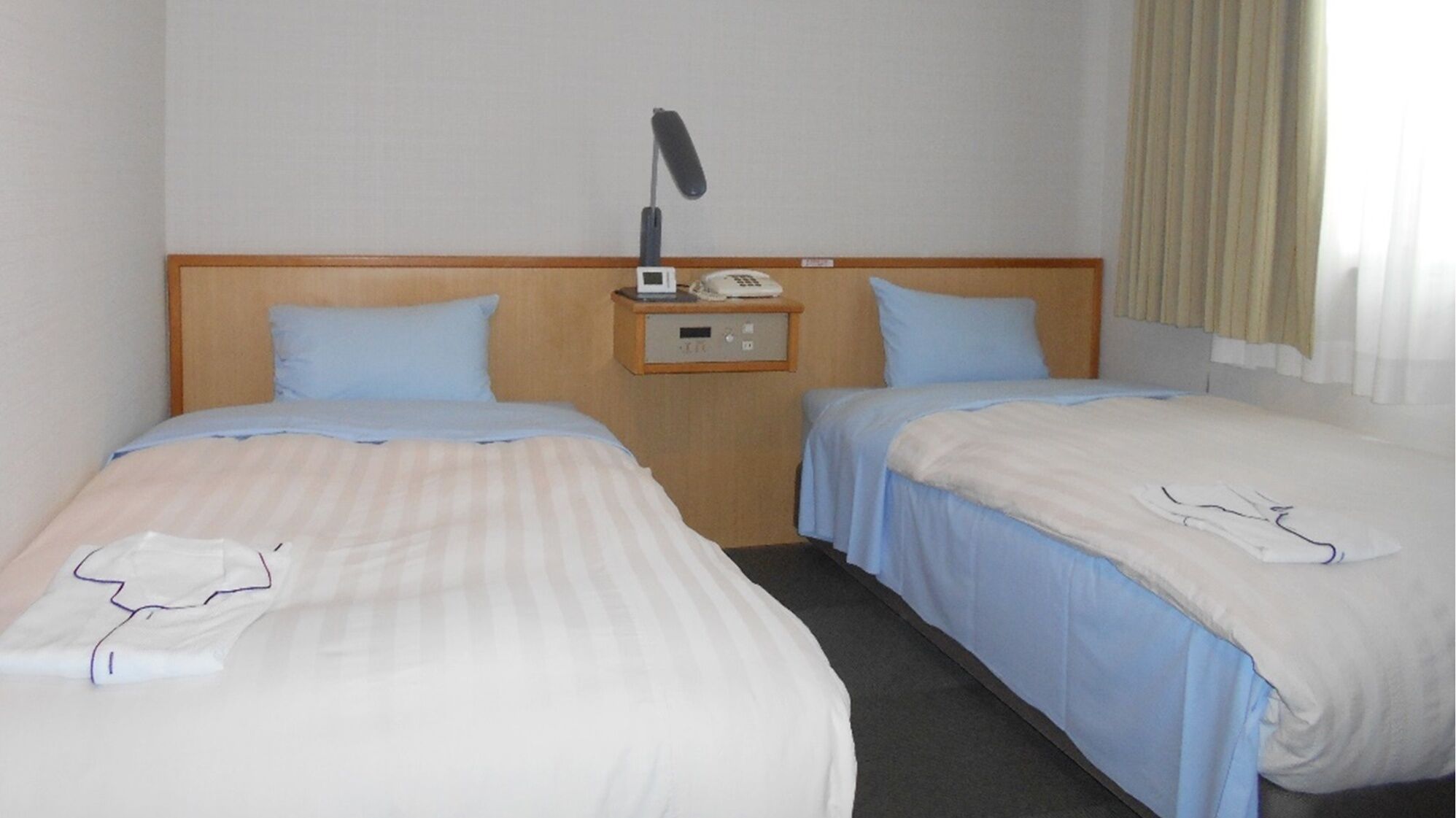A hotel room with two beds, a nightstand with a phone and a lamp, and a window with curtains.