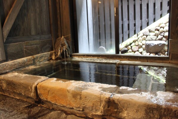 A stone-lined hot spring with a wooden fence and a waterfall in the background.