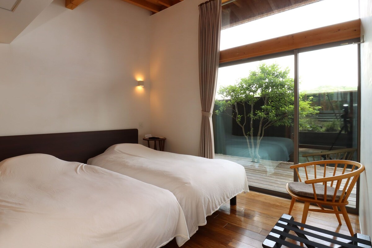 A hotel room with two beds, a small table, a chair, and a large window with a view of greenery.