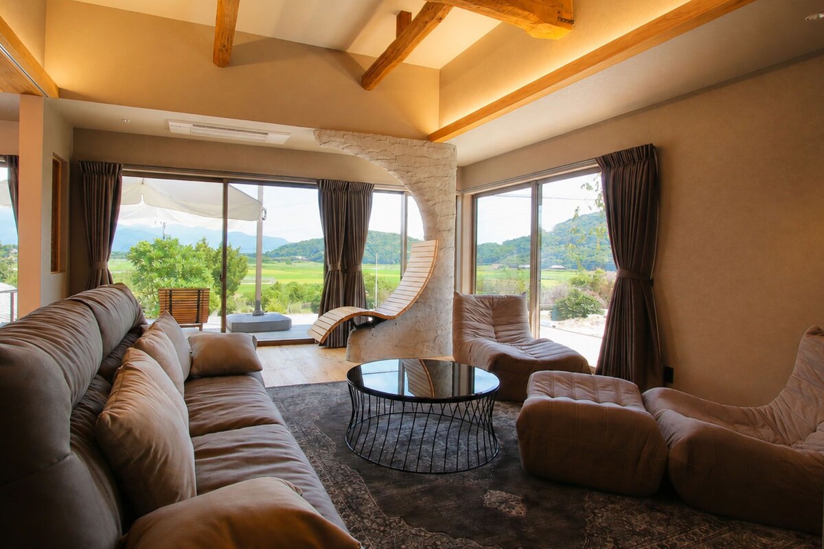 A spacious living room with a large sofa, a round coffee table, and a view of a lush landscape through large windows.