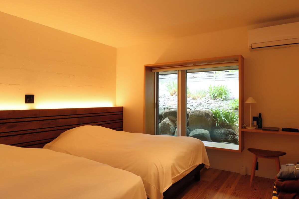 A hotel room with two beds, a window with a view of rocks and plants, a bedside table with a lamp, and an air conditioning unit on the wall.