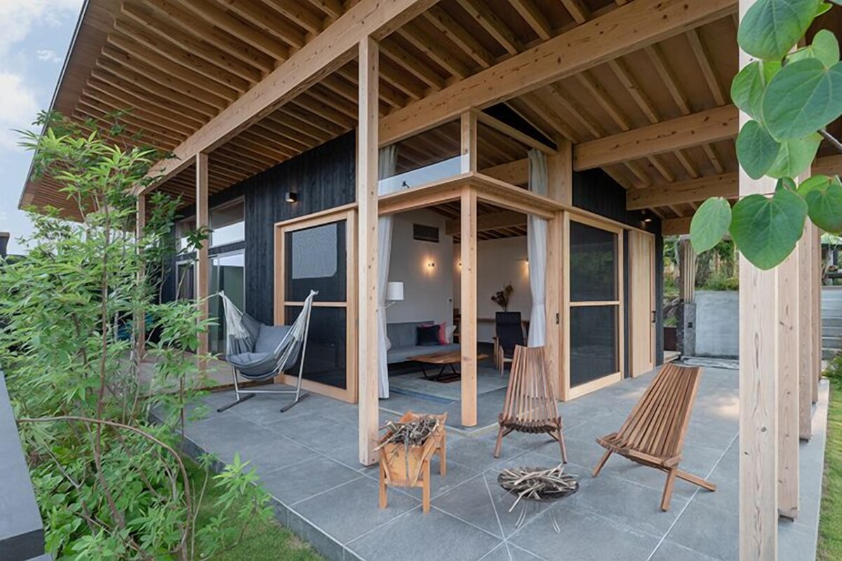 Un área de estar al aire libre con un diseño moderno, que incluye una hamaca, sillas de madera y un banco.