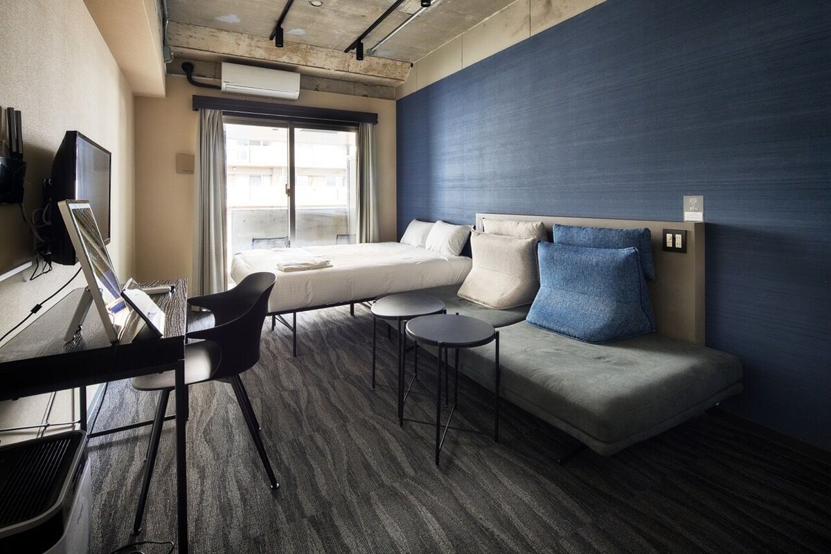 A hotel room with a bed, a desk with a chair, a small table, and a blue accent wall.