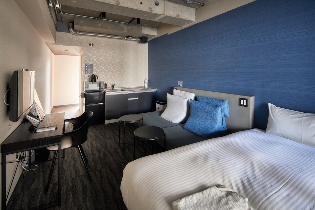 A modern hotel room with a bed, desk, chair, and kitchenette.