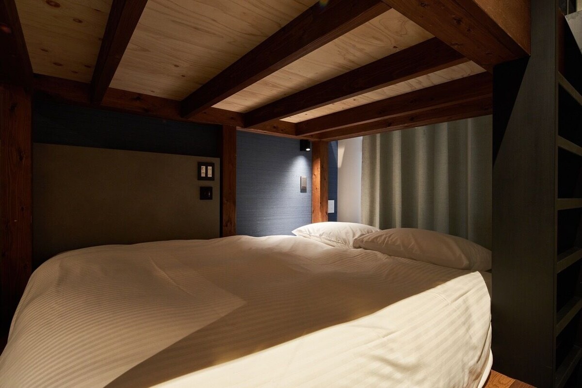 A bedroom with a large bed, wooden beams, a window with curtains, and a wall-mounted light.
